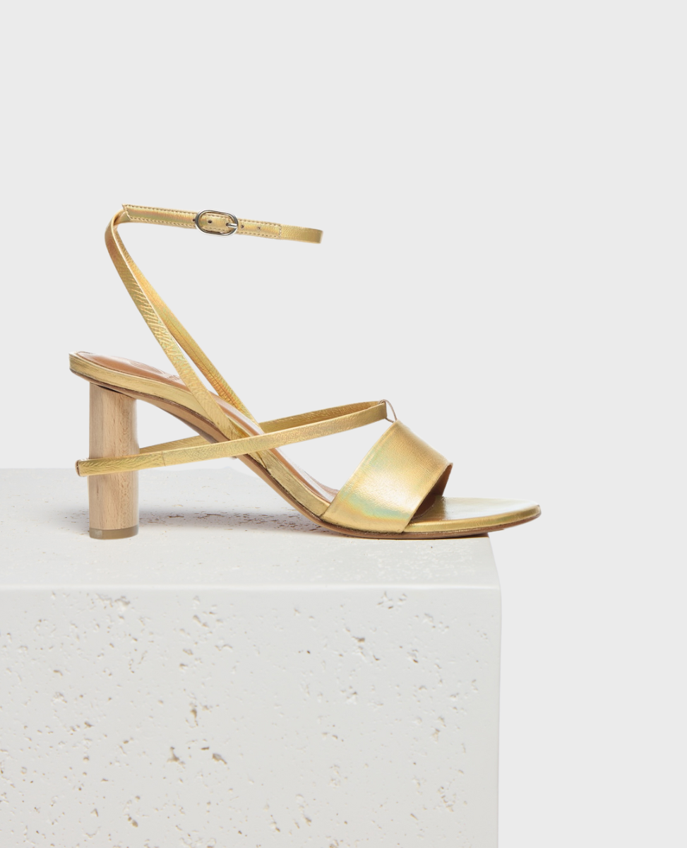 A gold sandal with a low, cylindrical wooden heel, featuring a sleek metallic finish, a thin ankle strap with a buckle, and a wide toe strap, all set on a minimalist white platform.
