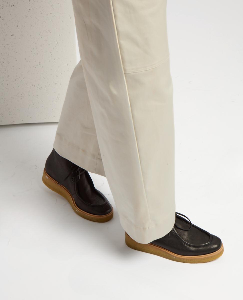 Black leather shoes with a smooth texture, featuring a moccasin-style toe and natural crepe soles, worn with light beige pants.