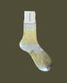 A single sock with a gradient design transitioning from light gold at the top to a silvery hue at the toe, featuring a smooth, slightly shiny texture and a rectangular label attached at the cuff. 3