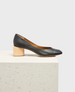 A sleek black leather pump with a rounded toe and a distinctive light wooden block heel, resting on a textured white surface. 1