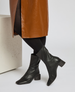 A pair of sleek, black, mid-calf leather boots with a square toe, low block heel, and a smooth, slightly shiny texture, worn with black tights and a brown textured skirt. 6