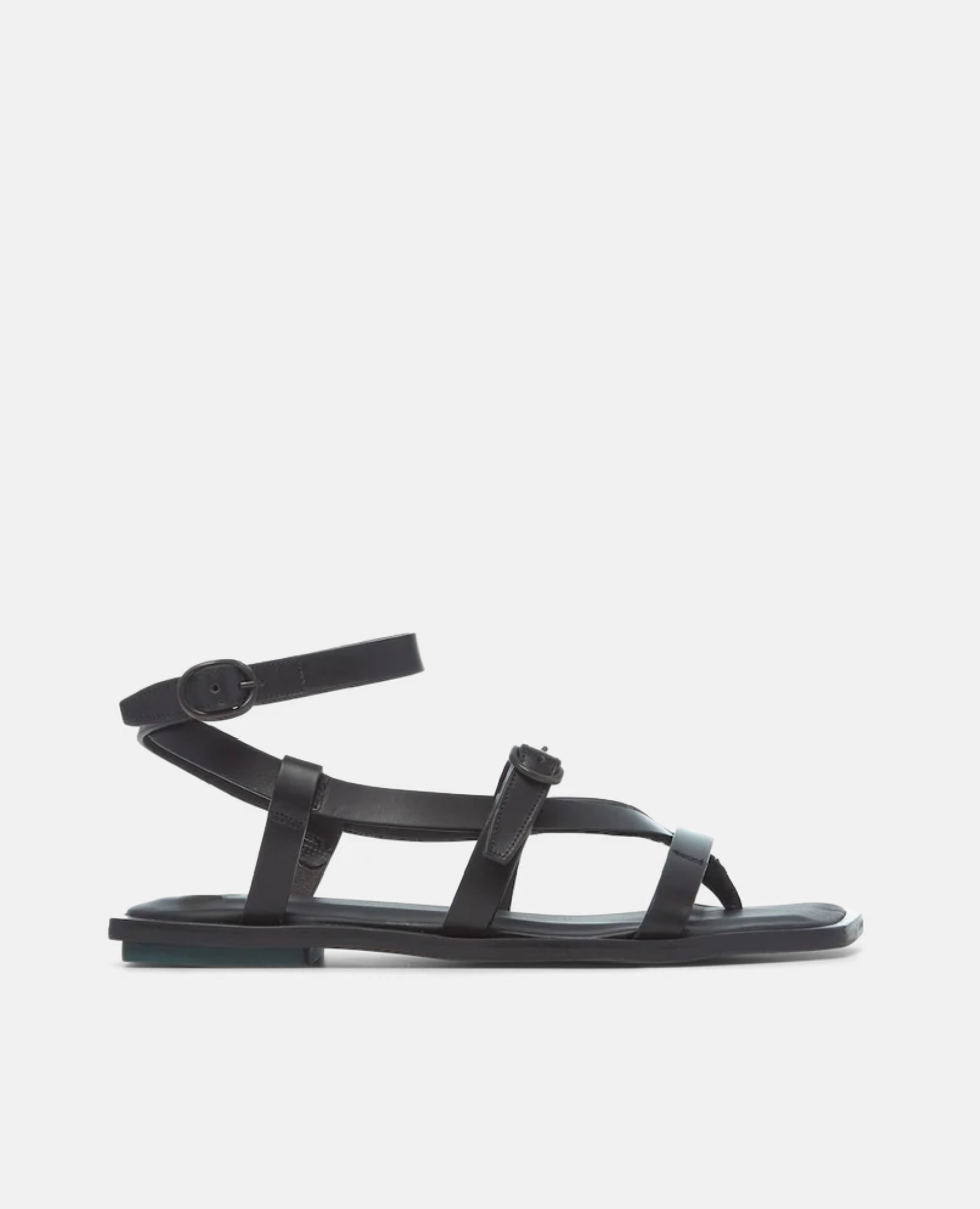 A sleek black sandal featuring a flat sole, multiple thin leather straps crossing over the foot, an adjustable ankle strap with a buckle, and a minimalist design.
