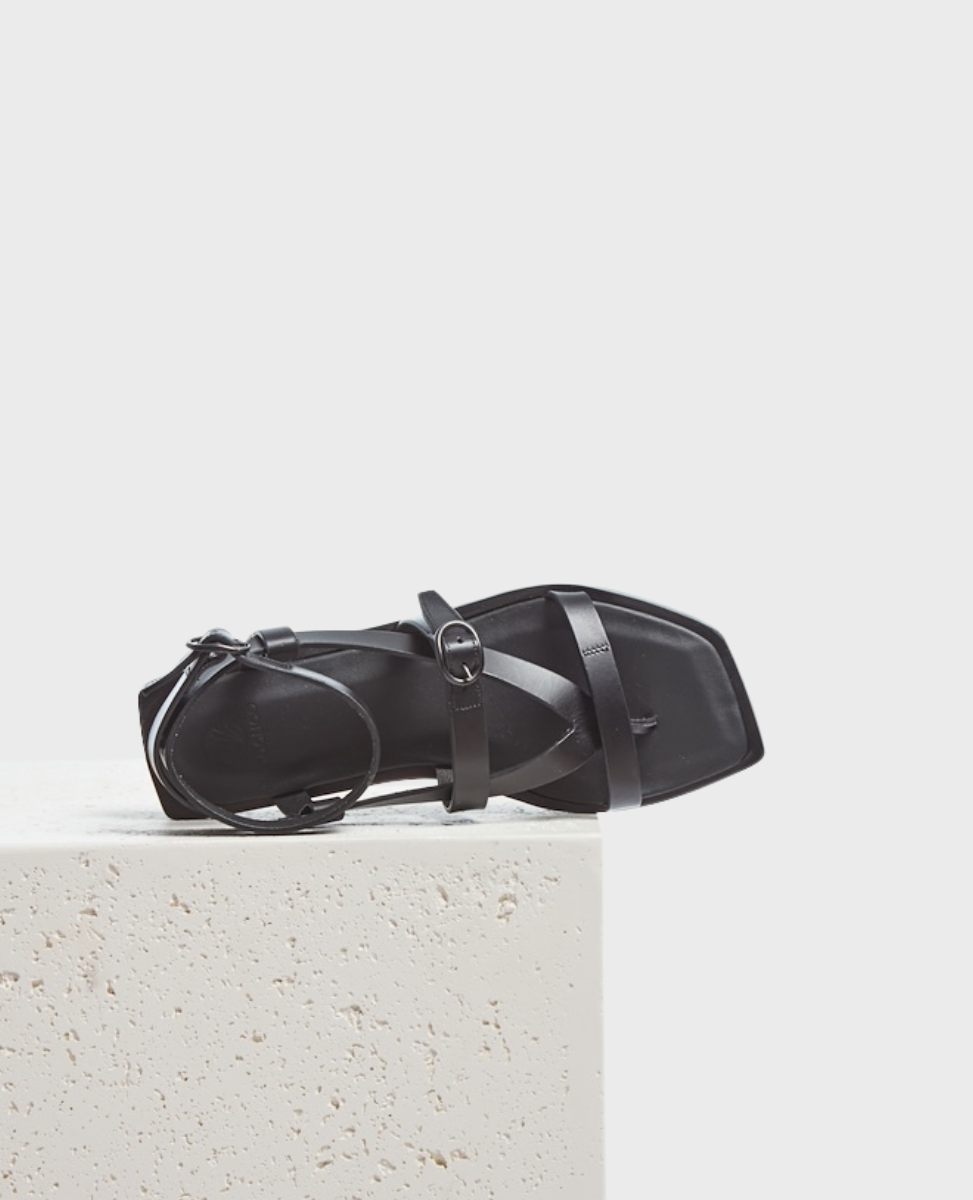 A sleek black sandal with a square toe, featuring thin, smooth leather straps and a small buckle, resting on a textured white surface.