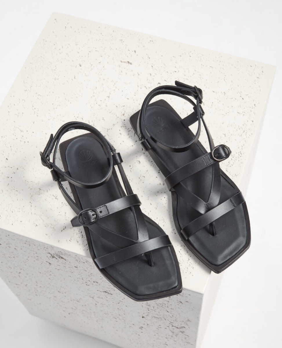 Black leather sandals with a square toe, featuring thin straps and adjustable buckles, resting on a textured white surface.