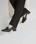 A pair of sleek, black leather loafers with a low, chunky heel, worn with black pants and light-colored socks, featuring a smooth texture and a minimalist design. 3