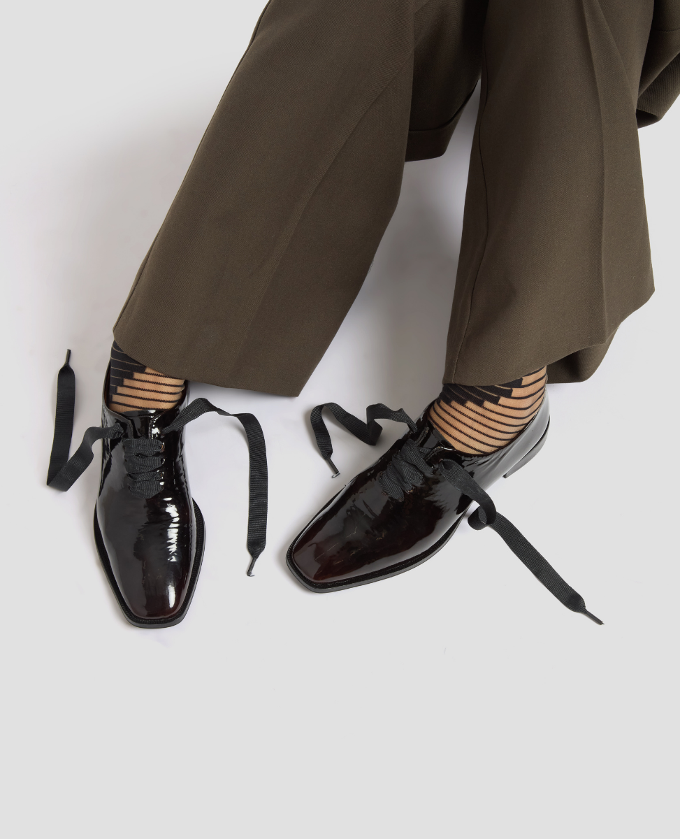Black patent leather shoes with bow details worn with brown pants on a white background