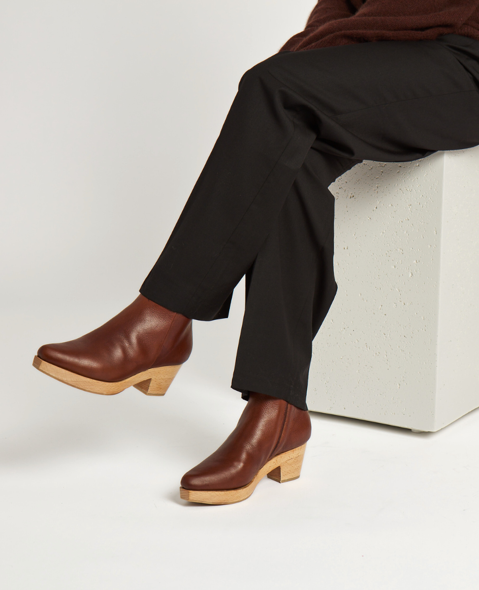 Brown leather clog boots with a smooth texture, featuring a wooden block heel and a slightly snipped toe, worn by a person sitting on a white block.