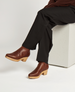 Brown leather clog boots with a smooth texture, featuring a wooden block heel and a slightly snipped toe, worn by a person sitting on a white block. 2