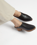 Black leather mule clogs with a smooth texture, featuring a wooden block heel and a shearling-lined interior, worn with light-colored pants. 4