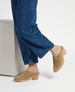 A pair of tan suede clogs with a wooden block heel, featuring a smooth, pointed toe and a slip-on design, worn with blue denim jeans. 3