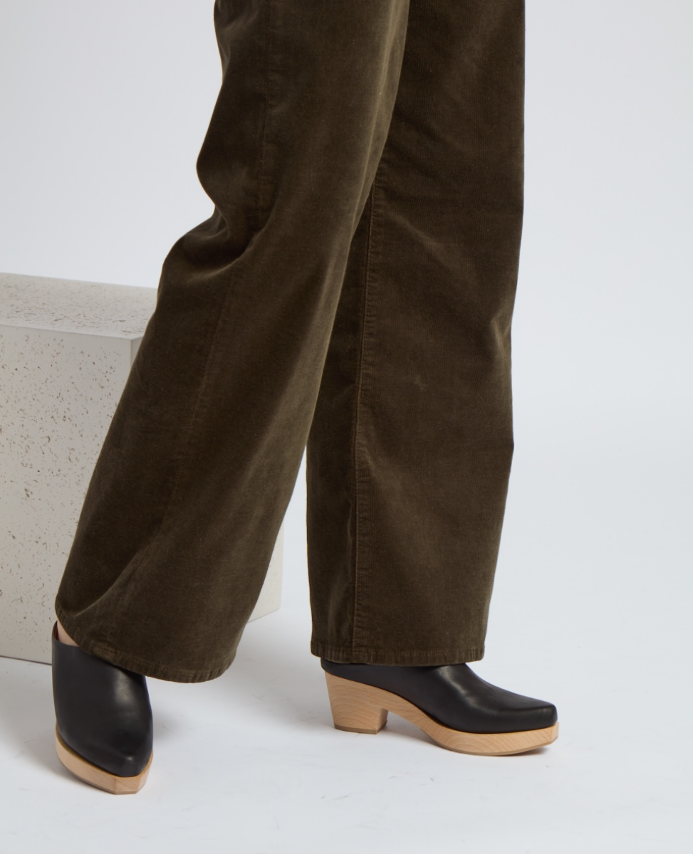Black leather clogs with a smooth finish, featuring a natural wood sole and a slightly elevated heel, worn with brown corduroy pants.