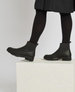 Black ankle boots with a smooth leather finish, round toe, elastic side panels, and a chunky rubber sole, worn with black tights and a black skirt. 2