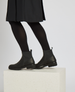 Black ankle boots with a smooth leather finish, elastic side panels, a pull tab at the back, and a low block heel, worn with black tights and a dark skirt. 4