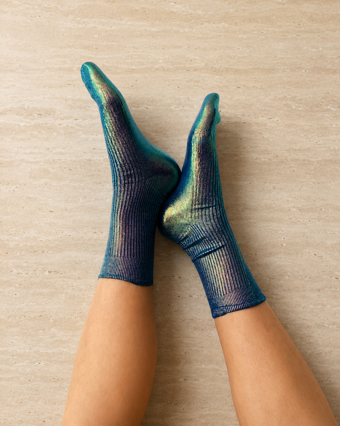 A pair of iridescent socks with a ribbed texture, displaying a gradient of blue, green, and purple hues, worn on feet against a stone
 background.