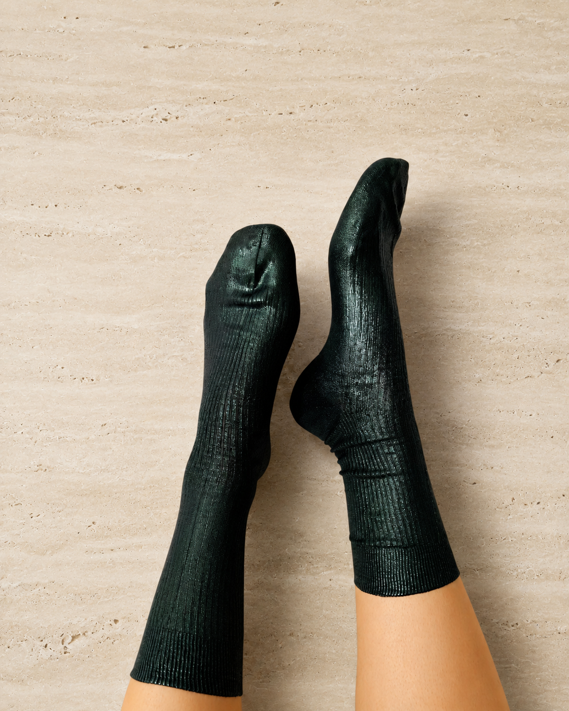 A pair of high-shine, metallic foil-laminated silk socks in dark green color, displayed on a person's legs against a stone background.