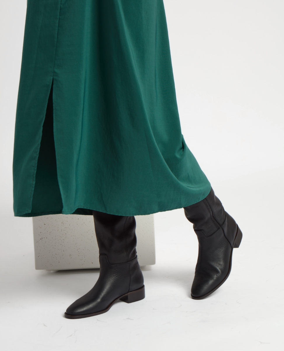 A pair of sleek black leather boots with a low heel and rounded toe, featuring a smooth texture and a slightly slouched shaft, worn with a long, flowing green skirt with a front slit.