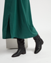 A pair of sleek black leather boots with a low heel and rounded toe, featuring a smooth texture and a slightly slouched shaft, worn with a long, flowing green skirt with a front slit. 3