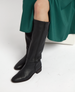 Tall black leather ridding boots with a smooth texture, featuring a side zipper and a low block heel, worn with a green dress. 4