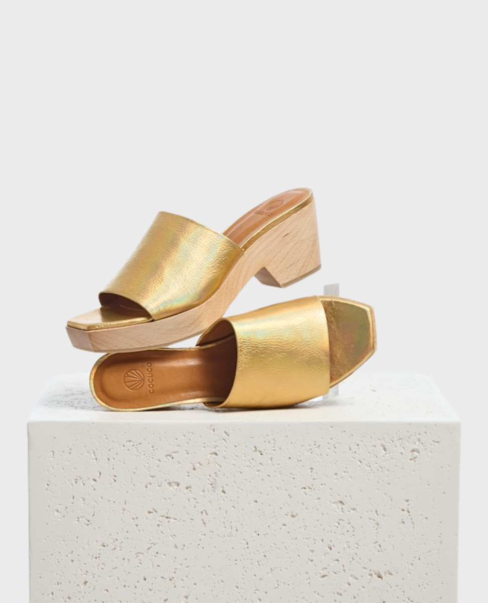 A pair of open-toe, gold iridescent slide sandals with a chunky wooden block heel and a square toe design, featuring a smooth, shiny texture and a minimalist style.