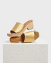 A pair of open-toe, gold iridescent slide sandals with a chunky wooden block heel and a square toe design, featuring a smooth, shiny texture and a minimalist style. 1