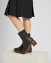 Black mid-calf leather boots with a smooth texture, featuring a chunky wooden heel and a slightly rounded toe, worn with a black skirt. 1