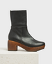 A black leather clog boot with a smooth texture, featuring a chunky wooden heel and platform, a side zipper, and a sleek, minimalist design. 2