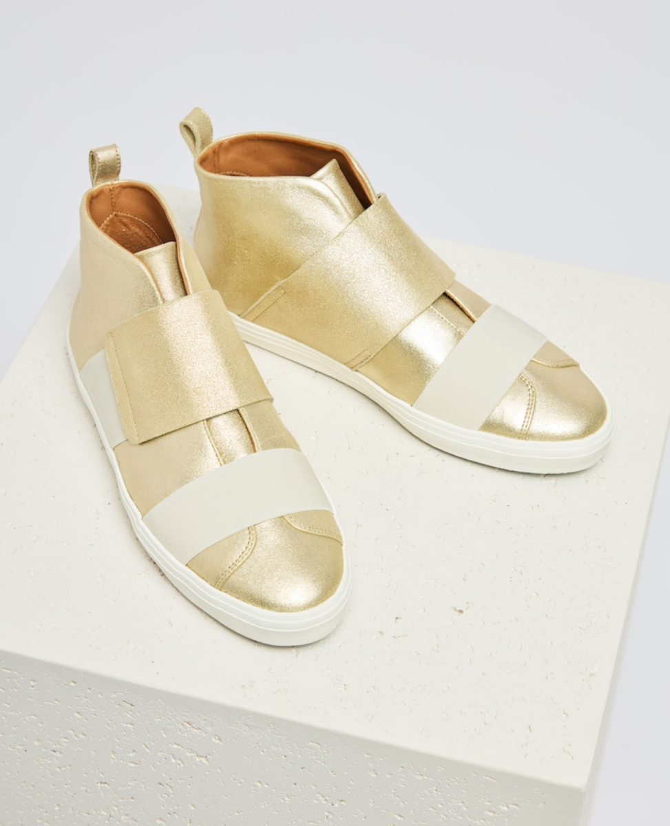 A pair of gold high-top sneakers with a metallic finish, featuring wide beige elastic straps across the top, white rubber soles, and a pull tab at the heel.