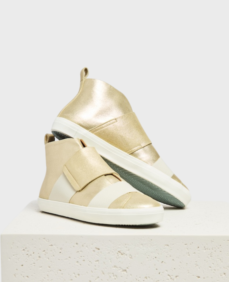 A pair of stylish high-top sneakers featuring a combination of metallic gold wide elastic straps, a white rubber sole, and a pull tab at the heel, displayed on a textured white pedestal.