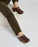 A pair of dark brown, metallic-finish clogs with a chunky wooden heel and a smooth, rounded toe, worn by a person in olive green pants. 3