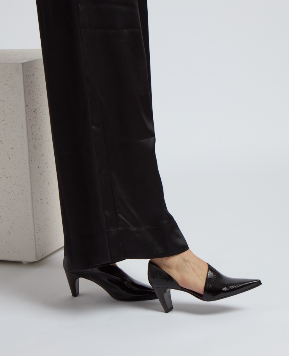 A sleek, black, pointed-toe shoe with a glossy finish and a mid-height, angular heel, worn with black pants.