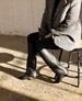 A person is seated on a stool wearing a gray, textured, herringbone-patterned coat, black pants, and black knee-high boots with a slight heel, against a backdrop of a white brick wall and a speckled concrete floor. 11