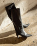 A pair of sleek, black, knee-high leather boots with pointed toes and short, angled heels, leaning against a textured white brick wall on a concrete floor. 10