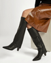 A pair of sleek, black knee-high boots with a pointed toe, block heel, and side zipper, worn with a shiny brown leather skirt. 4