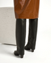 A pair of sleek, black, knee-high leather boots with a side zipper and a low, chunky heel, worn by a person in a glossy brown skirt, standing next to a textured, off-white block. 5