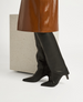 A pair of sleek, black leather knee-high boots with pointed toes and low, angled heels, featuring a glossy brown textured skirt in the background. 1