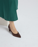 A brown, pointed-toe, mid-heel shoe with a smooth leather texture is worn on a foot, partially visible beneath a flowing green fabric. 2