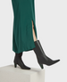 A pair of sleek, black leather ankle boots with pointed toes and mid-height, angular heels, worn with a long, green dress featuring a side slit. 4