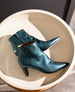 A pair of teal, pointed-toe ankle boots with side zippers is displayed on a round, cream-colored surface. 7