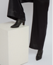 A pair of sleek, black leather ankle boots with pointed toes and chunky heels, worn with wide-legged black pants, is positioned on a textured, off-white rectangular pedestal. 6