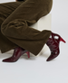 A pair of glossy, dark red, pointed-toe ankle boots with a mid-height stiletto heel, worn by a person in brown corduroy pants, resting on a white block. 3