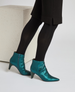 Teal, pointed-toe ankle boots with a glossy finish, side zippers, and low, tapered heels, worn with black tights and a black skirt. 3