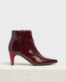 A sleek, dark red, patent leather ankle boot with a pointed toe, a side zipper, and a matching red high heel, displayed on a textured, off-white pedestal. 2