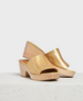 A pair of open-toe, iridescent gold clogs with a smooth leather upper and light wooden block heels, resting on a textured white surface. 1