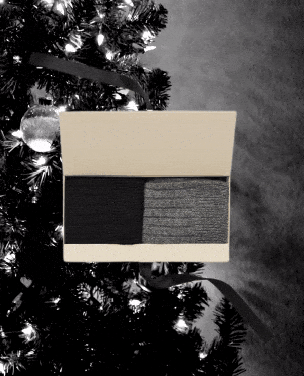 A rectangular beige gift box contains two pairs of socks, one solid black and the other with a subtle striped pattern, displayed against a backdrop of a decorated Christmas tree with lights.