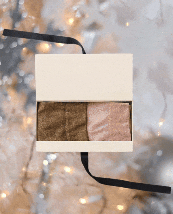 A rectangular cream-colored box with a black ribbon contains two folded silk scarves, one in shimmering gold and the other in soft pink, set against a blurred background of warm, twinkling lights.