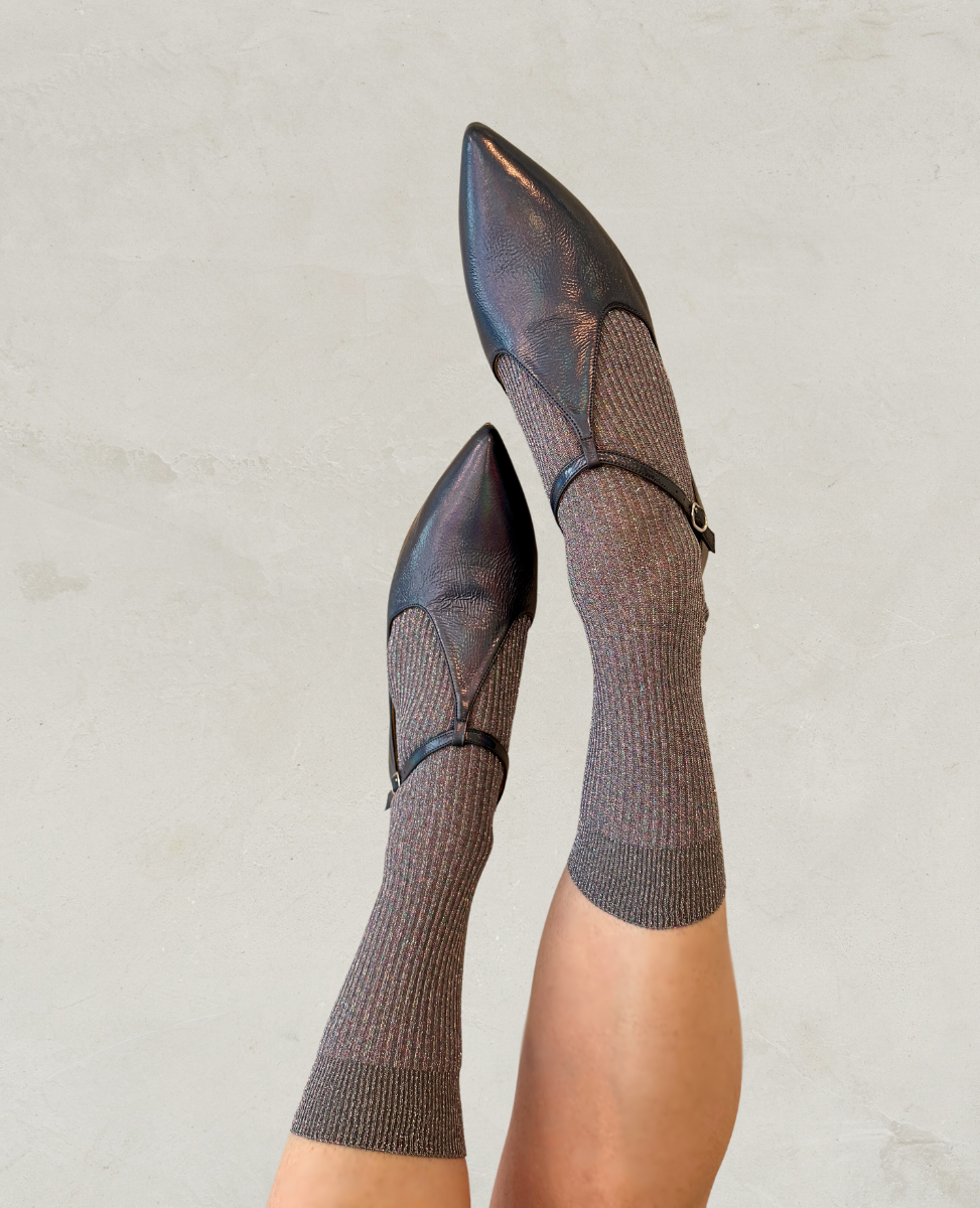 Pointed-toe black leather flats with a subtle sheen, featuring a thin strap with a small buckle, paired with ribbed gray socks.