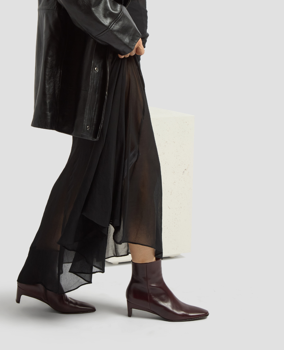 Person wearing a black coat, sheer black skirt, and brown ankle boots on a light gray background