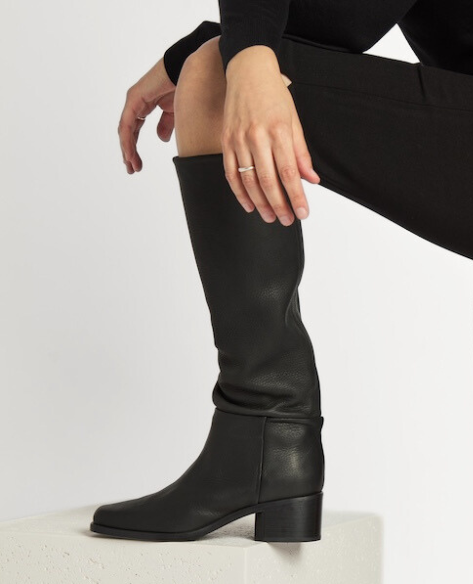 A sleek, knee-high black leather boot with a smooth texture, featuring a rounded toe, a low block heel, and a subtle seam running vertically along the side.