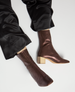 Shiny, metallic brown ankle boots with a pointed toe, smooth texture, and a contrasting light wooden block heel, worn with black satin pants. 1