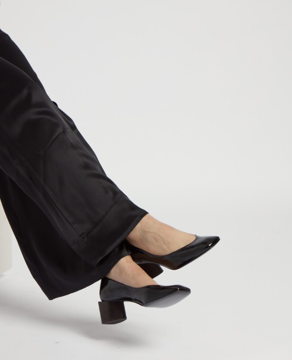 A pair of sleek, black patent leather heels with a pointed toe and a low, chunky heel, worn with black pants.